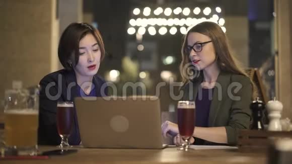两个带笔记本电脑的年轻女子晚上在户外咖啡馆聊天视频的预览图