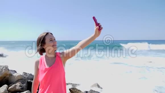年轻漂亮的白种人女孩带着手机站在岩石海滩上拍自拍照片海浪拍打着岩石视频的预览图