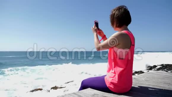 年轻美丽的高加索女人坐在岩石海滩上拿着电话拍照视频的预览图