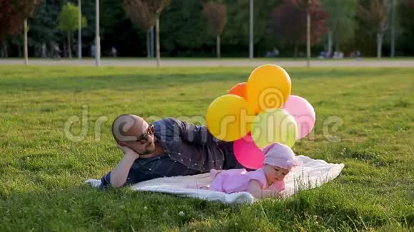 父亲拿着气球躺在白色的毯子上女儿躺在他身边在晴天在城市公园里采摘青草视频的预览图