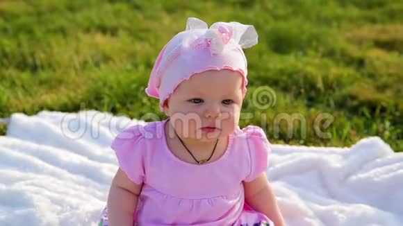 可爱的穿着粉色衣服的漂亮女婴紧张地坐在城市公园绿草上的白色毯子上视频的预览图