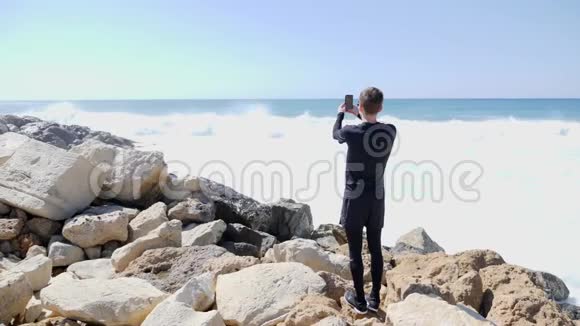 在一个阳光明媚的风日里年轻的帅哥在拍海洋和海浪拍打岩石海滩的照片溅起水花视频的预览图