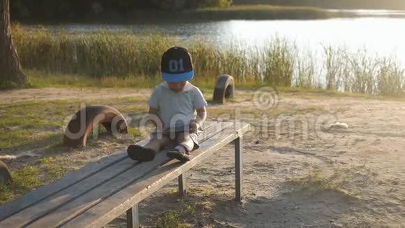 孤独的小男孩坐在长凳上手里拿着手机在夕阳下网络游戏生活方式户外视频的预览图