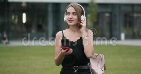 年轻女性在耳机里听音乐在城市散步时使用智能手机环顾四周视频的预览图