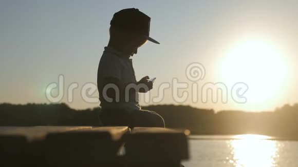 孤独的小男孩坐在长凳上手里拿着手机在夕阳下网络游戏生活方式户外视频的预览图