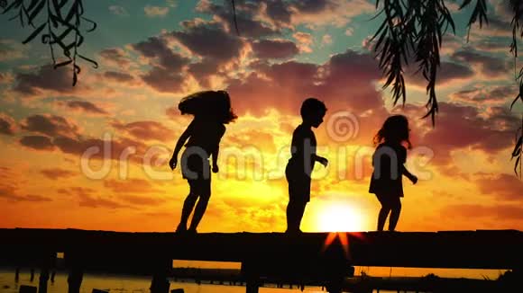 夕阳和欢乐的孩子们在湖水之上的木墩上跳跃的剪影视频的预览图
