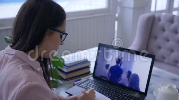 在线培训有魅力的年轻女子戴着眼镜在笔记本电脑上看视频时用笔写在笔记本电脑上视频的预览图