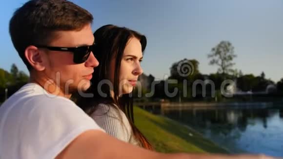 在池塘附近的绿色夏季公园里的浪漫情侣年轻一对微笑的夫妇在阳光公园聊天拥抱糖果花视频的预览图