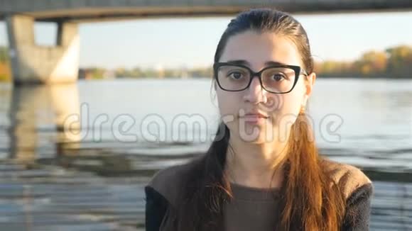 戴眼镜的漂亮女孩正看着相机微笑着可爱的微笑在河流的背景下视频的预览图