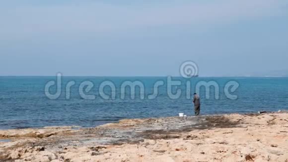 孤独的白种人渔夫站在岩石海岸上把鱼竿扔进海里老人在海上捕鱼视频的预览图