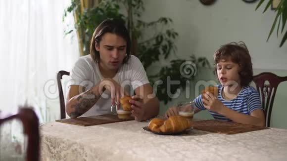 两个兄弟大人和小男孩坐在厨房的桌子上说话吃着加卡布奇诺的牛角面包兄弟会视频的预览图