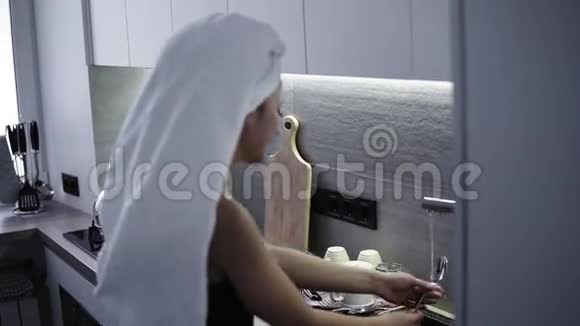 年轻女子头上戴白色浴巾在厨房洗碗穿着家居服的年轻女子早上视频的预览图