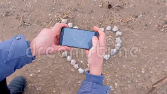 男人用手机双手特写在沙滩上用贝壳拍心视频的预览图