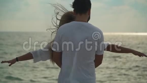 可爱的年轻夫妇穿着白色连衣裙站在海滩上靠近男人从背后搂着女人女人举手视频的预览图