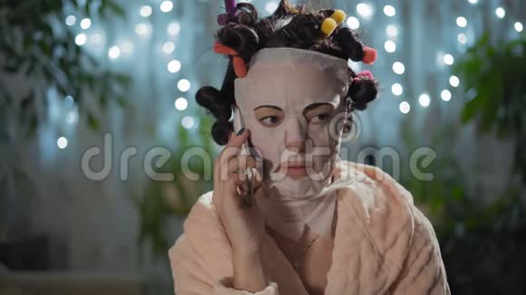 特写美丽的女人在一个令人耳目一新的面具与卷曲在电话上坐在房间里视频的预览图