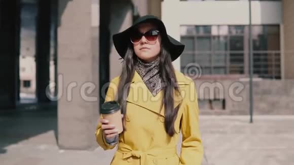 戴着黄色雨衣帽子和眼镜的时髦女孩走在街上四处张望喝着咖啡视频的预览图