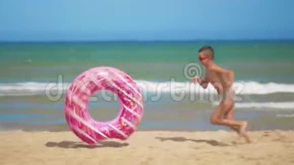男孩带着一个粉红色的充气甜甜圈沿着海滩跑把它卷在沙滩上贴在大海的背景下视频的预览图