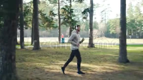 穿着运动服和运动鞋在公园里慢跑和通过现代音乐听音乐的留胡子年轻人的多利镜头视频的预览图
