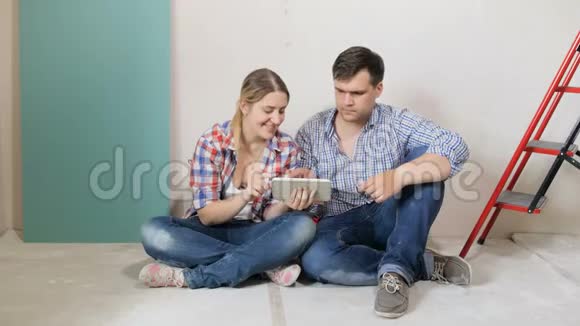 一对年轻夫妇坐在正在装修的新房子的地板上用互联网选择家具视频的预览图