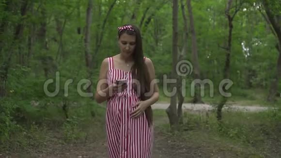 一个怀孕的女孩穿过公园一个穿着条纹白红色连衣裙的女孩通过电话说话抚摸着她的肚子视频的预览图