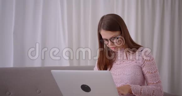 年轻漂亮的白种人女商人戴着眼镜在笔记本电脑上打字看着镜头微笑视频的预览图