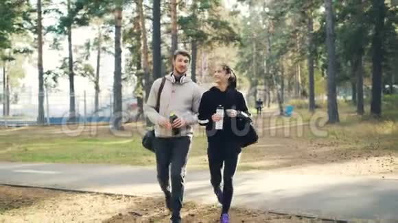 快乐的女孩和男孩穿着运动服手里拿着水瓶和袋子在城市公园里一起散步视频的预览图