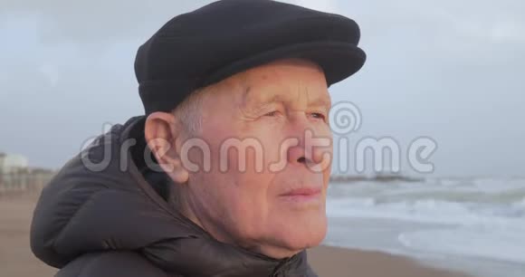 一位穿着平底帽和棕色外套的老人站在海滩上的画像视频的预览图