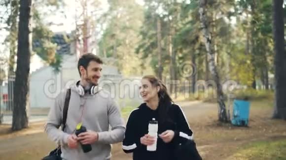 戴着耳机的年轻帅哥和他的女朋友聊天笑着拿着运动袋在公园里散步视频的预览图