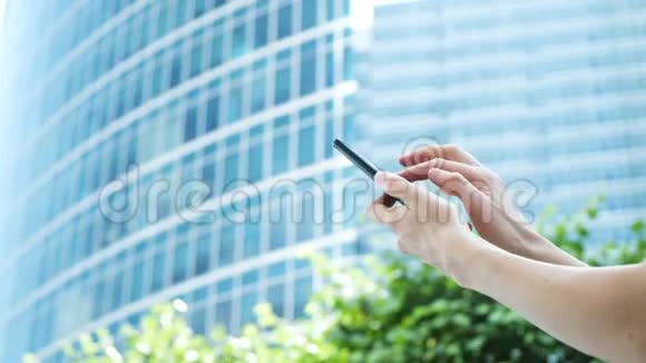 女人手在玻璃商务中心和绿树的背景下使用智能手机视频的预览图