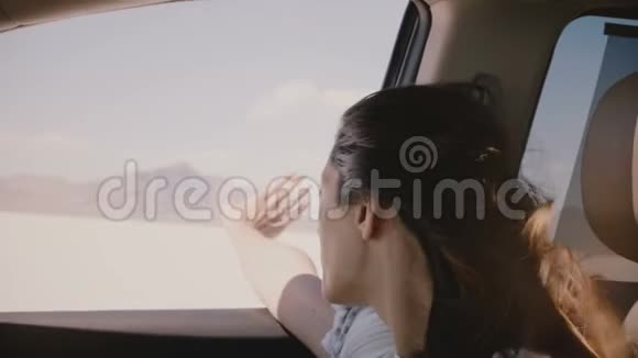 在盐湖沙漠一个快乐的年轻女人她的头发飞扬手从窗外开着车快速地移动着视频的预览图