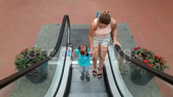 美丽的年轻母亲和蹒跚学步的儿子坐在购物中心的自动扶梯上的4k视频一个正在移动栏杆的男孩视频的预览图