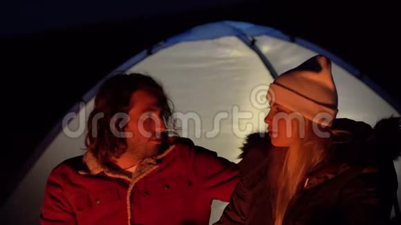 拍摄了一对年轻夫妇坐在火边聊天的特写镜头在海边用帐篷露营春天视频的预览图