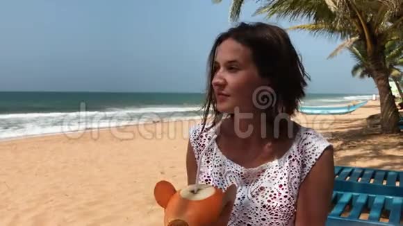 年轻有魅力的女士坐在海边热带海滩上度假喝椰子鸡尾酒视频的预览图