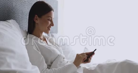 快乐的年轻女士早上躺在床上用电话视频的预览图