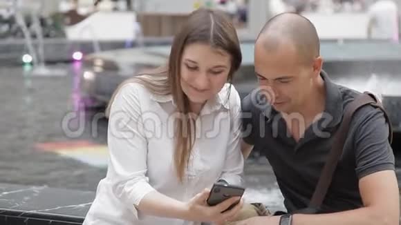 在喷泉背景下坐在购物中心的男人和女孩在电话里讨论照片笑了起来视频的预览图
