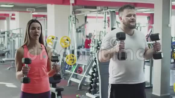 有个人教练的肥胖男人在健身房里用哑铃做身体抽搐超重的男性和女性教练燃烧视频的预览图