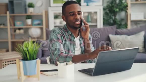 美国黑人自营职业者在家拿着笔记本电脑坐在家里用手机说话视频的预览图