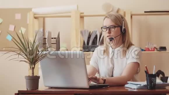 一个穿着白色上衣眼镜和耳机的女孩坐在一张桌子旁拿着笔记本电脑打字和与顾客交谈视频的预览图