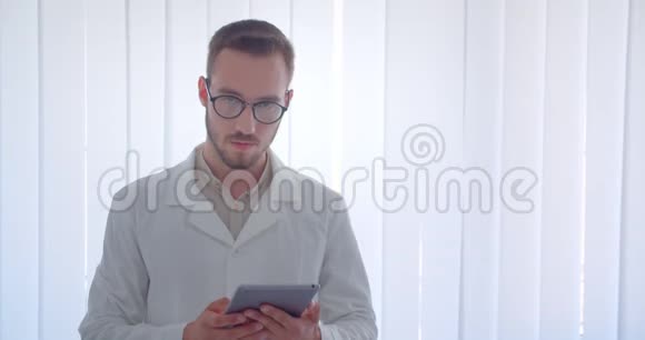年轻英俊的白种人男医生在白色公寓里拿着平板电脑看着镜头的特写画像视频的预览图