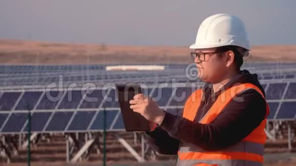 身穿橙色背心头戴安全帽手持太阳能电池板站在场边工作的亚洲人形象工程师视频的预览图