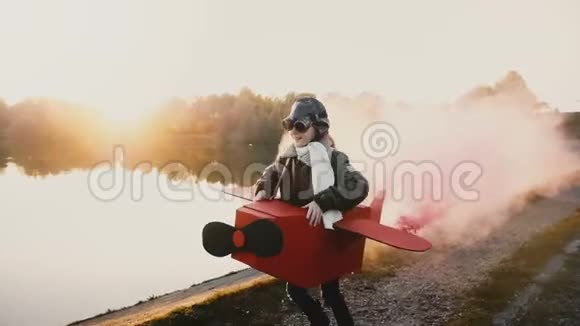 快乐的小飞行员女孩玩耍沿着日落湖跑步穿着有趣的纸板飞机服装带着彩色烟雾慢动作视频的预览图