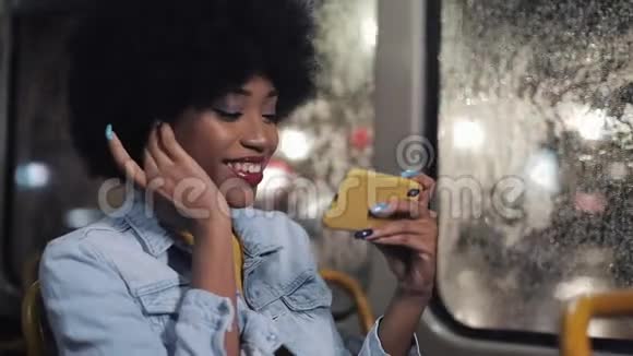 微笑着年轻的非洲裔美国妇女看有趣的视频智能手机骑在公共交通夜间视频的预览图