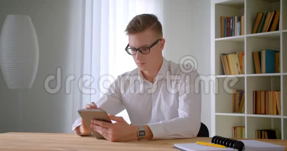 年轻英俊的白种人商人戴着眼镜用平板电脑在室内看着摄像机视频的预览图