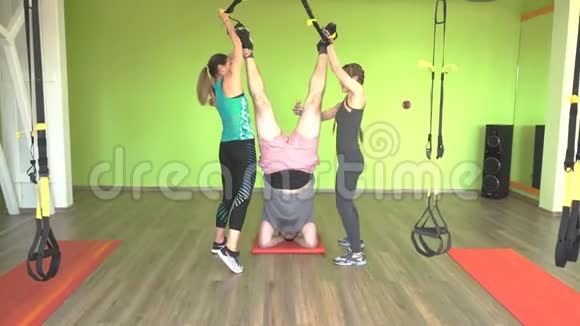 两个漂亮的白种人女孩教练帮助一个胖子在健身房锻炼铰链TRX视频的预览图