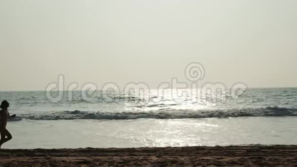 赤脚运动女孩跑步剪影沿着海浪游泳池跑步保持身体健康日落黑沙滩视频的预览图