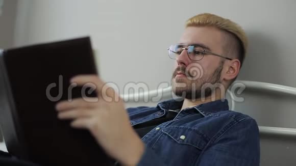 满意的年轻帅哥戴眼镜黄头发躺在床上看书的特写照片视频的预览图