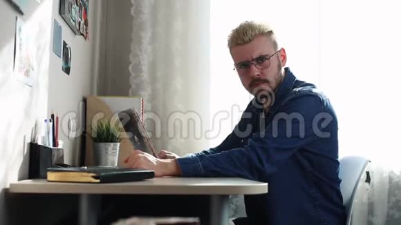 一个疯狂的家庭自由职业者带着眼镜和黄色头发在家工作使用笔记本电脑伤心失望的男人视频的预览图