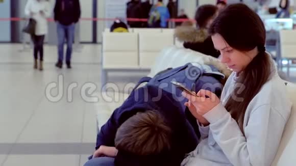 一名妇女带着疲惫的儿子在机场大厅里手里拿着一部手机等待航班侧视视频的预览图