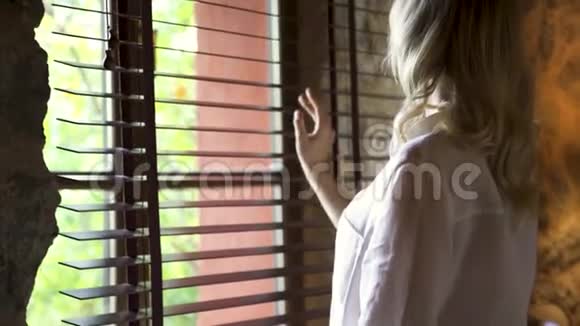 迷人的金发女郎用手触摸百叶窗望着玻璃窗外艺术年轻模特视频的预览图