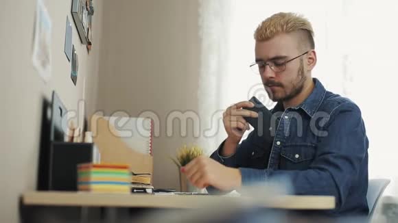 年轻的白种人在客厅里坐在桌子上时使用智能手机快乐的男人用电话打字视频的预览图